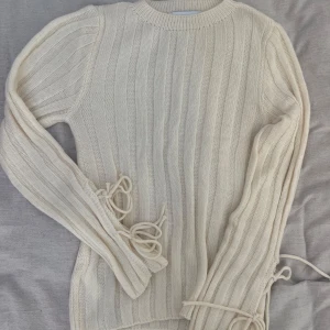 Vit ribbad stickad tröja med snörning - Säljer en vit stickad tröja med ribbat mönster och snygga snörningsdetaljer på ärmarna. Tröjan har rund halsringning och långa ärmar. Pris går att diskutera.