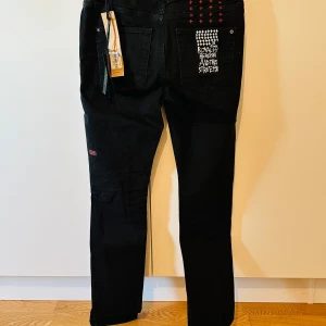 Svarta jeans Ksubi Chitch Laid Black - Svarta jeans från Ksubi, modell Chitch Laid Black. Snygga broderade röd plus på bakfickan och patch med texten 'Royalty Heroism And The Streets'. Klassisk femficksmodell med rak passform och unika detaljer.
