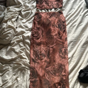 Brun two piece med rosor och matchande topp - Tvådelad klänning i glansig rosa satin med stora rosor som mönster. Lång kjol med rak passform och matchande bandeau-topp. Perfekt för dig som vill sticka ut med ett unikt set.