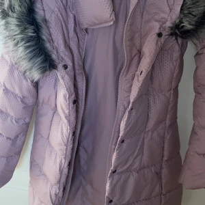 Ljuslila dunjacka med fuskpäls från The North Face - Säljer en ljuslila dunjacka från The North Face med avtagbar fuskpäls på huvan. Jackan har dragkedja och tryckknappar framtill samt quiltad design. Perfekt för kyliga dagar.