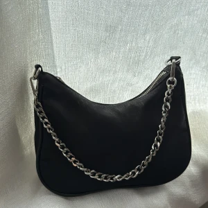 Svart axelväska med kedja - Snygg svart axelväska med silvrig kedja som axelrem och dragkedja upptill. Väskan har en mjukt rundad form och minimalistisk design, perfekt för att lyfta din outfit med en cool detalj. Passar till dig som gillar stilrena accessoarer.