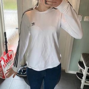 Vit långärmad tröja från Adidas - Säljer en vit långärmad tröja från Adidas med svarta ränder längs ärmarna och Adidas-logga på bröstet. Klassisk och sportig stil, perfekt till jeans eller träningsbyxor.