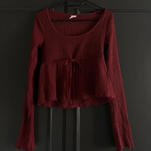 Vinröd långärmad topp med knyt - Säljer en vinröd långärmad topp med rund halsringning och knyt i midjan. Toppen har en lätt volang nedtill och är tillverkad i ett ribbat material som ger en snygg struktur. Kommer inte till användning och är endast använd max 3 gånger 