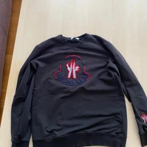 Svart sweatshirt från Moncler med broderad logga - Säljer en svart sweatshirt från Moncler i storlek L. Tröjan har en stor broderad Moncler-logga på bröstet i rött, vitt och blått samt en mindre brodyr på ärmen. Klassisk rund halsringning och ribbade muddar.