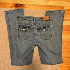 Blå bootcut jeans med fickdetaljer - Säljer ett par klassiska blå jeans med bootcut passform. Jeansen har två bakfickor med lock och knappdetaljer samt en ljus tvätt. Perfekta för dig som gillar en tidlös stil med lite utsvängda ben. Från Gina tricot Young 