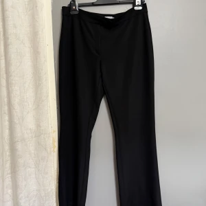 Svarta kostymbyxor från Nelly - Svarta kostymbyxor från Nelly med klassisk rak passform. Byxorna har hög midja och är tillverkade i ett mjukt, slätt tyg som ger en stilren look. Perfekta att matcha med en skjorta eller stickad tröja.