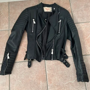 Svart skinnjacka från Voyelles - Snygg svart skinnjacka från Voyelles med coola detaljer som dragkedjor, bälte i midjan och quiltade partier på axlar och ärmar. Jackan har lång ärm och flera fickor med dragkedja. Perfekt för en edgy look.