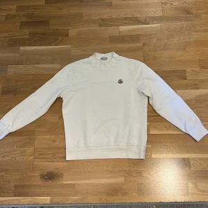 Vit sweatshirt från Moncler - Säljer en stilren vit sweatshirt från Moncler med rund hals och broderad Moncler-logga på bröstet. Tröjan har långa ärmar och är tillverkad i mjukt material som känns skönt mot huden. Perfekt för en clean och klassisk look.