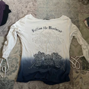 Vit och blå långärmad topp med snörning och tryck - Säljer en långärmad topp i bomull med coolt tryck och texten 'Follow the Westwind' framtill. Tröjan har snygg blå ombre-effekt nertill och snörningsdetaljer längs ärmarna. Perfekt för dig som gillar unika detaljer och streetstyle.