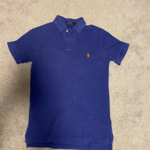 Blå piké t-shirt från Polo Ralph Lauren - Säljer en klassisk blå piké t-shirt från Polo Ralph Lauren med orange broderad logga på bröstet. T-shirten har krage och knappar framtill. Perfekt för en stilren look.