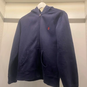 Mörkblå hoodie från Ralph Lauren - Mörkblå hoodie med dragkedja från Ralph Lauren. Klassisk design med röd broderad logga på bröstet och känguruficka framtill. Perfekt för en avslappnad stil och enkel att matcha med det mesta.