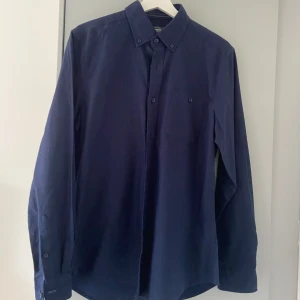 Mörkblå skjorta från Dressmann - Snygg mörkblå skjorta från Dressmann med klassisk krage och knappar framtill. Skjortan har lång ärm och en slimmad passform. Skjortan har knappt använts och är i bra skick. Storlek: M