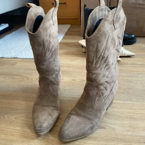 Beige mocka cowboy boots - Säljer ett par snygga beige cowboy boots i mocka med klassiska broderade detaljer och rund tå. Skorna har dragflikar upptill för enkel påtagning och en låg klack. Perfekta för dig som vill ha en trendig westernstil.