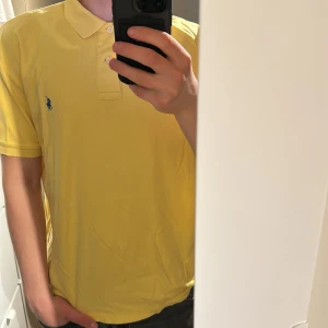 Gul pikétröja från Ralph Lauren - Säljer en klassisk gul pikétröja från Ralph Lauren med blå logga broderad på bröstet. Tröjan har korta ärmar, krage och knappar framtill. Perfekt för en stilren och avslappnad look. Gamla taggen 