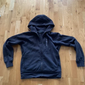 Svart hoodie från Sail Racing - Svart hoodie från Sail Racing med dragkedja framtill och mysigt fleecefoder på insidan. Tröjan har huva och två fickor samt diskret logga på bröstet. Perfekt för kyliga dagar.