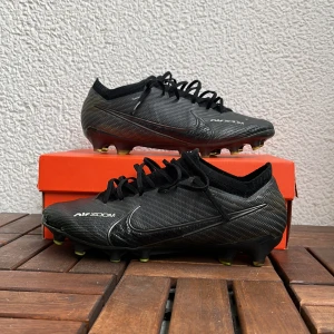 Svarta Nike Air Zoom fotbollsskor - Säljer ett par svarta Nike Air Zoom elite fotbollsskor med snörning och rund tå. Skorna har en lätt och sportig design med mönstrad ovandel och gula detaljer på sulan. Perfekta för dig som vill ha bra grepp på planen. Skorna har använts ungefär 3 månader och är lite smått skadade på sidorna av skorna. Vi kan också förhandla om priset! Box ingår!