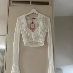 Vit spetsig croppad blus med volang - Säljer en vit croppad blus från edikted i spets med långa ärmar och volangdetaljer längs den v-ringade urringningen💗💗 aldrig använd med lappen kvar!!