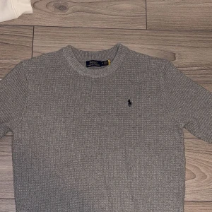 Grå stickad tröja från Polo Ralph Lauren - Säljer en klassisk grå stickad tröja från Polo Ralph Lauren med rund halsringning och den ikoniska broderade loggan på bröstet. Perfekt för lager-på-lager och enkel att matcha med det mesta.