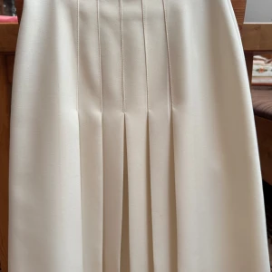 Vintage 80-tal  retro vit kjol, storlek 34 Fabric by Klopman - Säljer en vit kjol from Fabric by Klopman i 100% polyester (Dacron). Kjolen har klassiska veck framtill och stängs med dragkedja och knapp bak. Perfekt för dig som gillar en stilren och tidlös look. Mest troligt att kjolen är från tidigt 1980-tal. Säljs i befintligt skick 