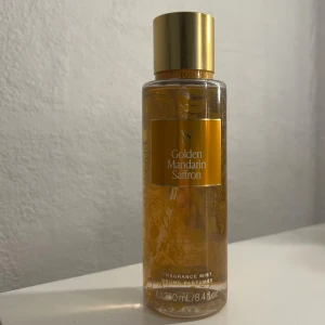 Golden Mandarin Saffron Fragrance Mist - En lyxig doftmist från Victoria's Secret med noter av gyllene mandarin och saffran. Flaskan är genomskinlig med guldiga detaljer och ett guldlock. Rymmer 250 ml och ger en fräsch och varm doftupplevelse.