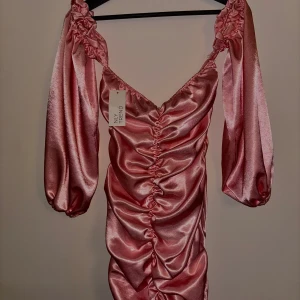 Rosa satin klänning från NLY Trend - Säljer en rosa satin klänning från NLY Trend i storlek 38. Klänningen har v-ringning, långa voluminösa ärmar och rynkade detaljer framtill. Perfekt för dig som vill sticka ut med en glansig och trendig look. Sitter tight. 