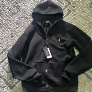 Svart hoodie från Polo Ralph Lauren - Svart hoodie från Polo Ralph Lauren med dragkedja framtill och klassisk logga broderad på bröstet. Tröjan har huva med snörning och två fickor framtill. Perfekt för en avslappnad stil.