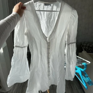 Vit klänning med knappar från Asos  - Säljer en vit klänning från asos med v-ringning och långa, lätt utsvängda ärmar. Klänningen har knappar framtill och är tillverkad i ett tunt, luftigt material. Perfekt för sommarens alla tillfällen! Första knappen är sönder annars är den fint skick 