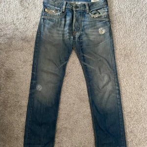 Diesel jeans - Säljer ett par blå jeansbyxor från Diesel Timmen med slitna detaljer och unika broderier på bakfickan. Byxorna har raka ben, klassisk femficksdesign och snygga slitningar framtill och baktill. Perfekta för en avslappnad stil.