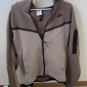 Beige och brun hoodiejacka från Nike - Nike hoodiejacka i beige och brunt med svarta detaljer och dragkedja framtill. Jackan har huva, lång ärm och en ficka med dragkedja på ena ärmen. Perfekt för en sportig och avslappnad stil. Denna färgen är både snygg och svår att hitta nu förtiden. 