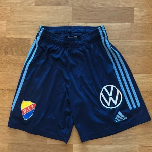 Två par blåa fotbollsshorts från Adidas med DIF-logga - Två par mörkblåa fotbollsshorts från Adidas med ljusblå ränder längs sidorna, DIF-emblem på ena benet och Volkswagen-logga samt Adidas-logga på andra benet. Resår i midjan och sportig design. Så två identiska par för 100kr sammanlagt