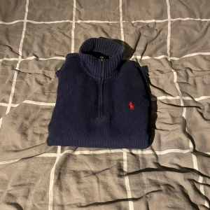 Marinblå stickad tröja med halv dragkedja från Polo Ralph Lauren - Säljer en marinblå stickad tröja från Polo Ralph Lauren med halv dragkedja och hög krage. Tröjan har lång ärm och den klassiska röda loggan broderad på bröstet. Perfekt för lager-på-lager och snygg till jeans.