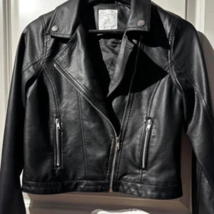 Svart skinnjacka med dragkedja - Snygg svart skinnjacka med klassisk bikerkrage och silverfärgade dragkedjor framtill. Jackan har långa ärmar och detaljerade sömmar längs axlar och ärmar. Perfekt för en cool och stilren look.