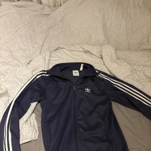 Blå Adidas track jacket med vita ränder - Klassisk blå Adidas track jacket med vita ränder längs ärmarna och dragkedja framtill. Jackan har hög krage och två fickor på sidorna. Perfekt för en sportig och avslappnad stil. Tröja med hög krage som kan vikas som gör de mycket snyggt
