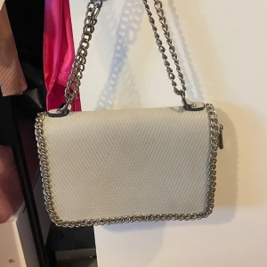 Beige axelväska med kedjedetaljer - Säljer en snygg beige axelväska med silverfärgad kedja som axelrem och runt kanterna. Väskan har ett diskret mönster, dragkedjeficka på baksidan och röd insida. Helt ny oanvänd 