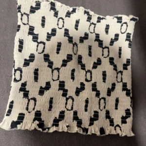Vit och svart smockad tubtopp med mönster - Säljer en smockad tubtopp i vitt med svart grafiskt mönster i storlek xs/s. Toppen är elastisk och har volangkant nertill och upptill. Perfekt att styla till jeans eller kjol för en trendig look. 