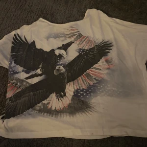 Vit t-shirt med örntryck från Gina Tricot - Vit t-shirt från Gina Tricot med stort tryck av två örnar i svart och vitt med amerikanska flagan. T-shirten har korta ärmar och en avslappnad passform. Perfekt för en cool och avslappnad stil.