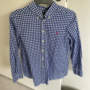 Blårutig skjorta från Ralph Lauren - Klassisk blårutig skjorta från Ralph Lauren med knappar framtill och button-down krage. Skjortan har lång ärm och en liten broderad logga i rött på bröstet. Perfekt för en stilren och tidlös look.