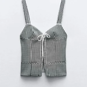 Svartvit rutig korsettopp med knytning - Trendig korsettopp i svartvitt rutigt mönster med breda axelband och söt knytning framtill. Toppen har figurnära passform och är perfekt för varma dagar.