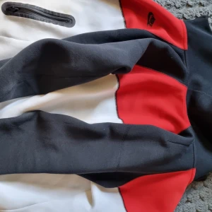 Nike zip hoodie i rött, vitt och svart - Nike hoodie med dragkedja i färgerna röd, vit och svart. Jackan har svarta detaljer på ärmar och axlar, samt två svarta dragkedjefickor framtill. Klassisk Nike-logga på bröstet och hög krage. 