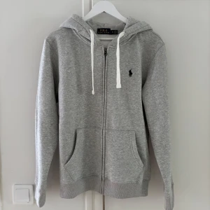 Grå zip hoodie från Polo Ralph Lauren - Säljer en klassisk grå zip hoodie från Polo Ralph Lauren med dragkedja och vit snörning i huvan. Tröjan har två fickor framtill och den ikoniska loggan broderad på bröstet. Perfekt för en avslappnad stil.
