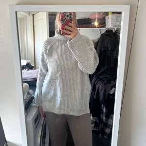 Ljusgrå stickad polotröja - Mysig ljusgrå stickad polotröja med ribbad struktur och långa ärmar. Tröjan har en avslappnad passform och hög krage, perfekt för kyliga dagar.