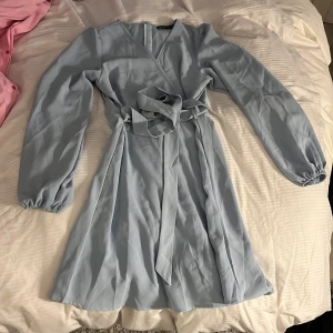 Ljusblå omlottklänning från SHEIN - Säljer en ljusblå omlottklänning från SHEIN med långa ärmar och knytband i midjan. Klänningen har en feminin siluett och är perfekt för vår och sommar. Enkel och stilren design som passar till många tillfällen.