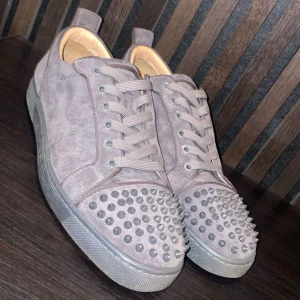  Christian Louboutin skor  - Säljer ett par gråa sneakers från Christian Louboutin med ikonisk röd sula och coola nitar på tån. Skorna har snörning och är tillverkade i mocka för en lyxig känsla. Perfekta för dig som vill sticka ut med din stil.