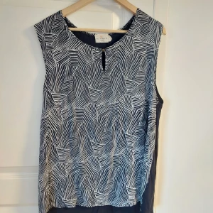 Svartvit ärmlös topp från Holly & Whyte - Säljer en svartvit ärmlös topp från Holly & Whyte by Lindex. Toppen har ett grafiskt mönster med vita linjer på svart botten och en liten knappdetalj vid halsen. Perfekt för en stilren och modern look.