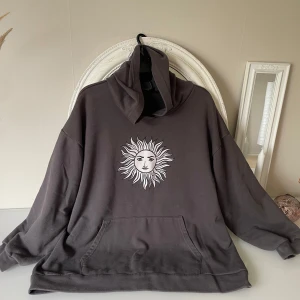Grå hoodie med soltryck från DAZY - Säljer en oversized grå hoodie från DAZY med en stor vit sol med ansikte tryckt på bröstet. Tröjan har huva och en stor magficka framtill. Perfekt för dig som gillar chill och unik stil.saknar band i luvan.