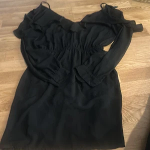 Svart offshoulder klänning från H&M Divided - Svart kortklänning från H&M Divided med offshoulder-design och långa ärmar. Klänningen har resår i midjan och tunna axelband. Perfekt för dig som gillar en stilren och trendig look.