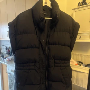 Svart dunväst från Gina Tricot - Säljer en svart dunväst från Gina Tricot med hög krage och dragkedja framtill. Västen har en puffig design och två fickor på sidorna. Perfekt att slänga över en hoodie eller tröja för extra värme.