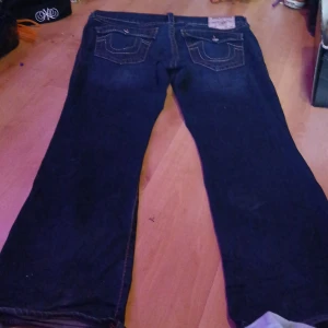 Mörkblå jeans från True Religion - Säljer ett par mörkblå jeans från True Religion med klassiska kontrastsömmar och stora bakfickor med lock och knapp. Jeansen har en rak och något vidare passform, perfekt för en avslappnad stil.