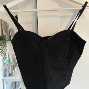 Svart korsettopp från H&M Divided - Snygg svart korsettopp från H&M Divided med smala axelband och markerade kupor. Toppen har en liten rosett framtill och är tillverkad i en stretchig viskosblandning som sitter tight och formar kroppen.