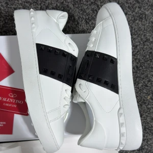 Valentino Garavani vita sneakers med svarta nitar - Säljer ett par vita Valentino Garavani sneakers med bred svart rem över vristen dekorerad med svarta nitar. Skorna har rund tå, platt sula och klassisk snörning. Märkeslogga på plösen och detaljerad sula. Perfekta för dig som gillar exklusiv streetstyle.